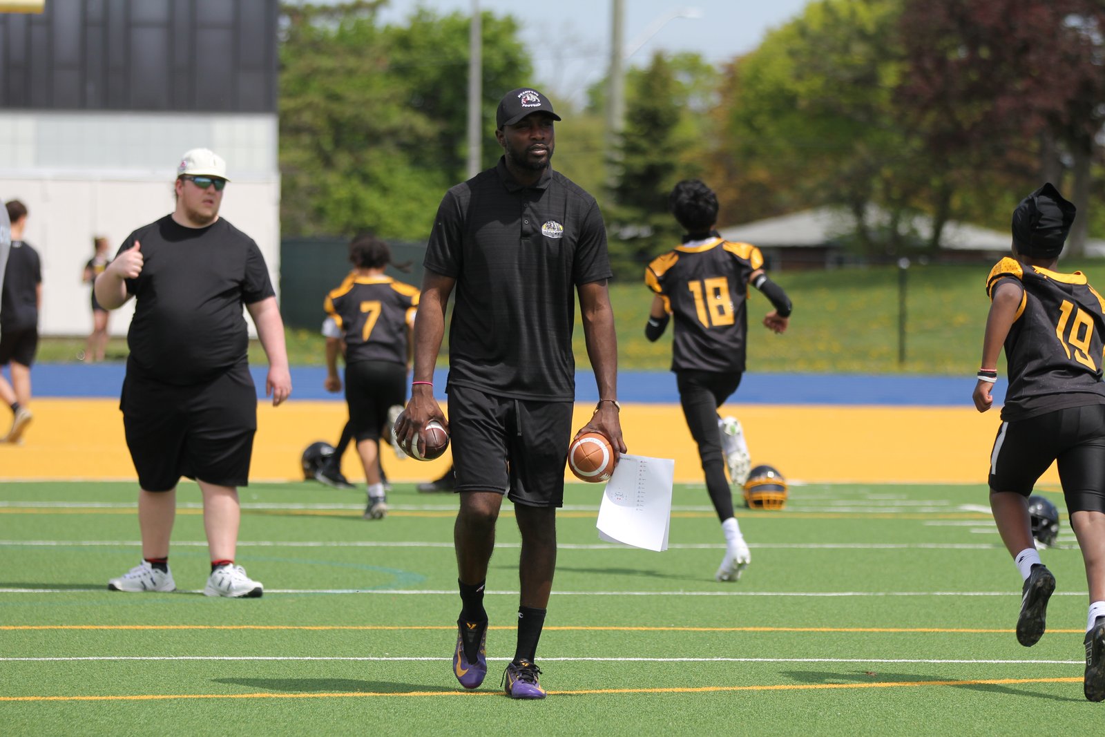 A Bulldogs Bantam coach walking onto the field at warm-up, footballs in hand, with players in the background