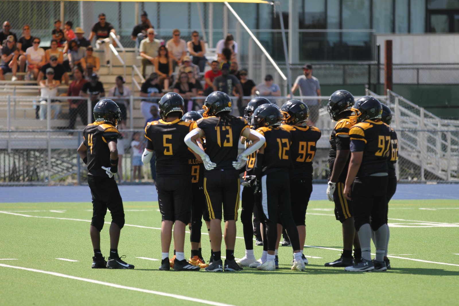 Bulldogs Bantam offense huddled at midfield with parents and fans in the stands behind them