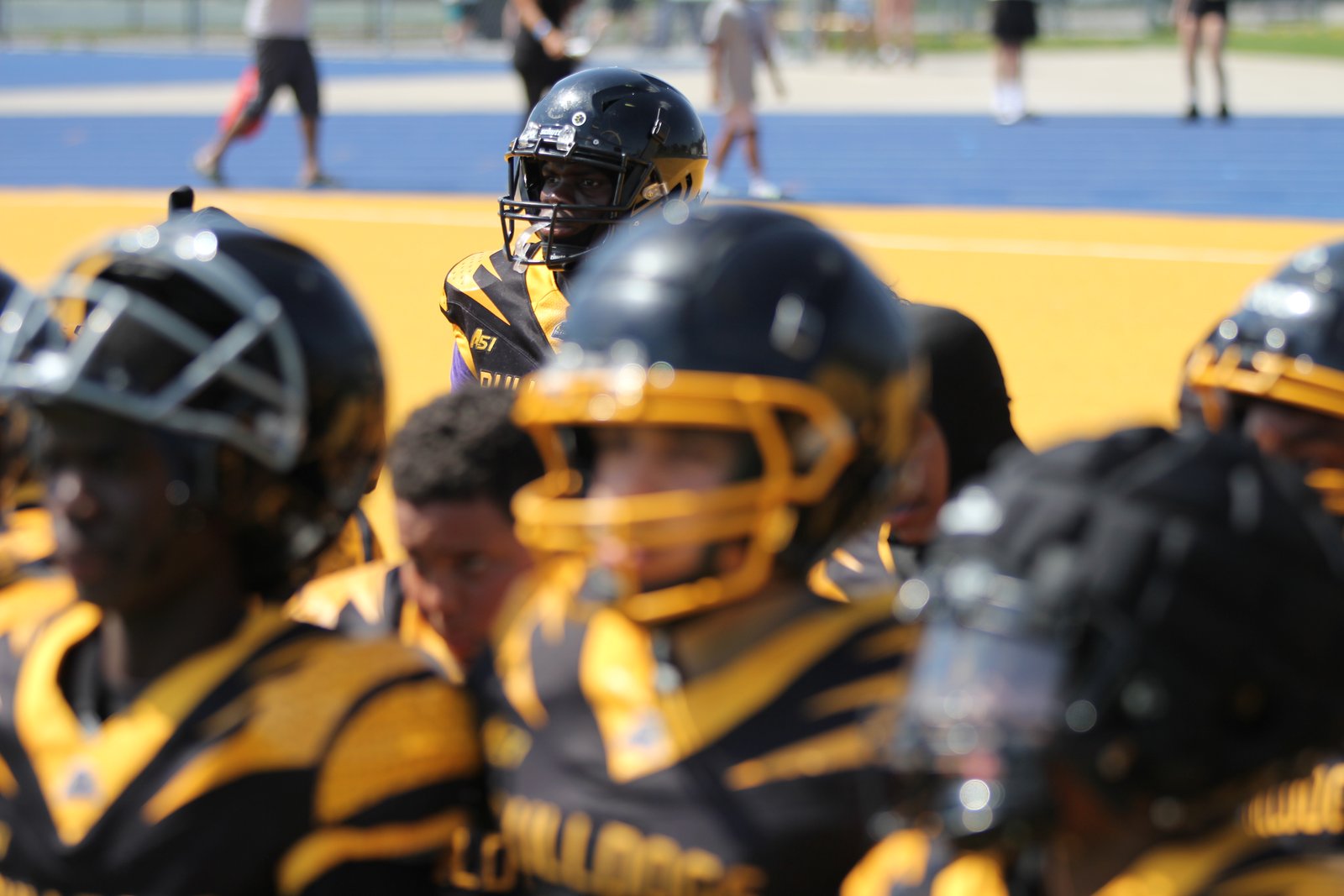 A Brampton Bulldogs U-14 Bantam player on the sideline, helmet on, looking through the facemask toward the camera while teammates fill the foreground in black and gold uniforms